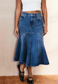 Denim buzzerskirt med høj talje, der har kontrastsyninger, lukning med knapper foran og en knælængde. Bæres med sorte flade sko.