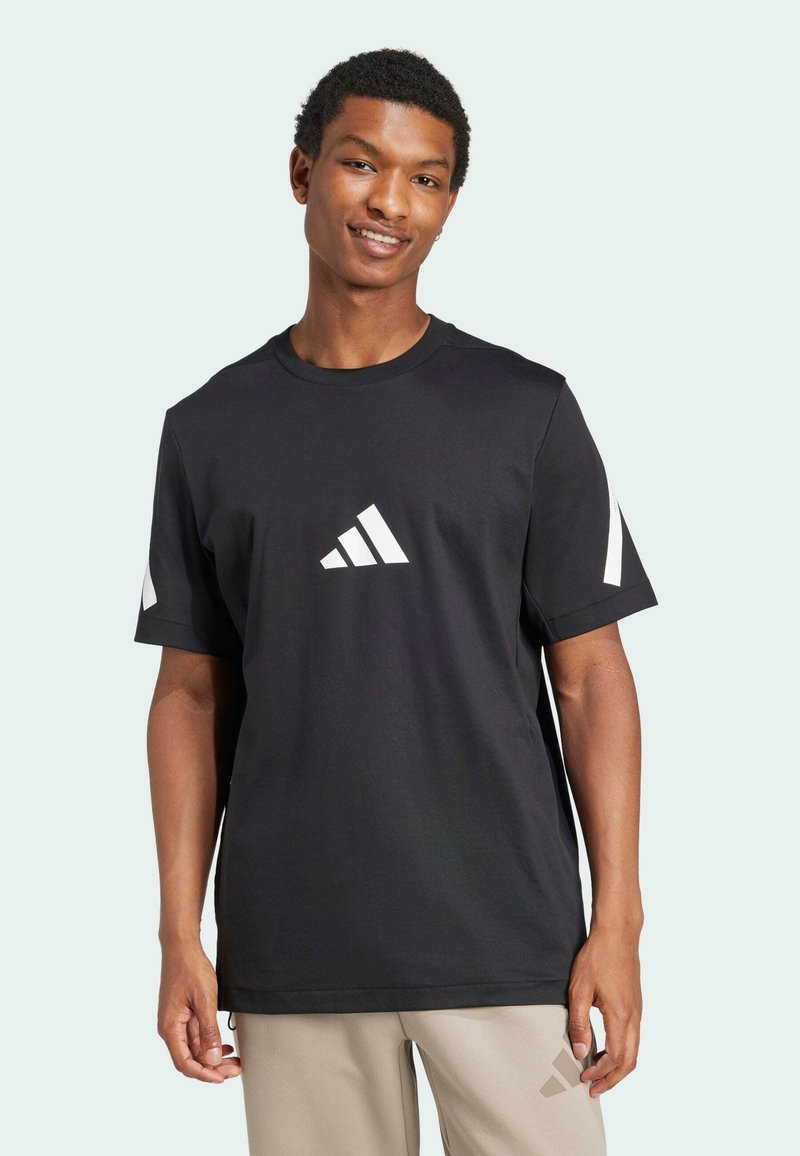 Giovane uomo con una maglietta nera Adidas con logo bianco sul petto e sulle maniche, sorridente con pantaloni beige Adidas su sfondo semplice.