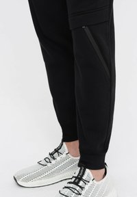 Schwarze Jogginghosen mit einem figurbetonten Design, ausgestattet mit einer Reißverschlusstasche und gerippten Bündchen, kombiniert mit leichten, perforierten weißen Sneakers.