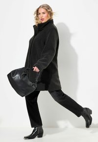 Cappotto in pile nero con colletto alto, di colore uniforme; abbinato a jeans neri e stivali alla caviglia spessi; tiene in mano una grande borsa nera.