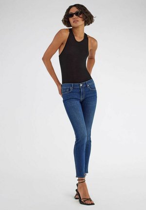 Frau trägt schwarzes ärmelloses Oberteil, blaue enge Jeans, schwarze Riemchenabsätze und schwarze Sonnenbrille, steht mit den Händen hinter dem Rücken.