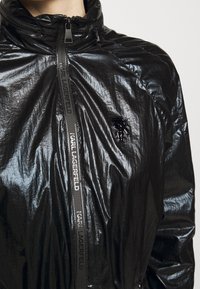 Czarna kurtka wykonana z błyszczącego, marszczonego materiału, z wysokim kołnierzem, zapinana na zamek błyskawiczny z przodu oraz markowym nylonowym suwakiem. Posiada naszywkę z logo na klatce piersiowej.