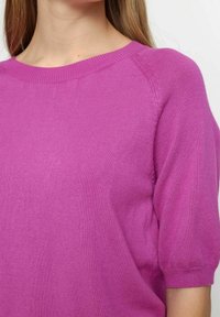 Pull ajusté à manches courtes en tissu maille rose vif. Caractérisé par un col rond côtelé et une texture lisse avec un léger éclat.