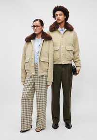 Beige, textúrált kabátok barna szőrme gallérral és szív alakú gombokkal. A modellen világoskék ing és kockás, bő szárú nadrág van.