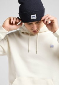 White hoodie with drawstrings and a front pocket, featuring a blue and white Lee logo patch. Worn with a dark blue ribbed beanie.