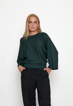 Mujer con cabello largo y rubio lleva un suéter suelto verde oscuro y pantalones negros, de pie con las manos en los bolsillos frente a un fondo blanco.