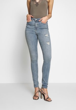 Kvinde i lyseblå højtaljede, revnede skinny jeans, olivengrøn tanktop med blonder og sorte højhælede sandaler med stropper, hænderne i lommerne.