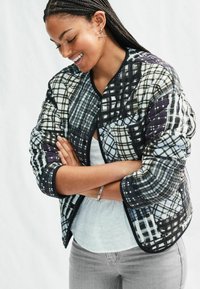 Lepaskan jaket bomber berlapis dengan corak plaid hitam, putih, dan ungu, dilengkapi dengan kerah dan manset bertepi. Dipadukan dengan atasan putih dan celana abu-abu.