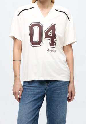 White short-sleeve shirt with large dark "04" and "MODSTRÖM" text, worn with blue jeans by a person with tattoos on both forearms.