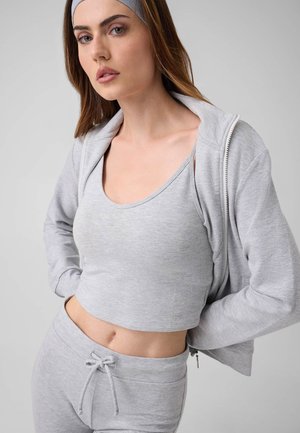 Frau mit hellgrauem Stirnband, Reißverschluss-Hoodie, Crop-Top und Jogginghose mit Kordelzug steht mit den Händen in der Hüfte vor einfachem Hintergrund.