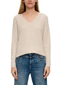 Donna che indossa un maglione a collo a V beige a maniche lunghe e jeans di denim blu, in piedi con le mani rilassate ai lati.