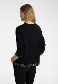 Suéter negro con cuello redondo, mangas largas con puños y dobladillo decorados, adornado con tachuelas circulares doradas y plateadas.