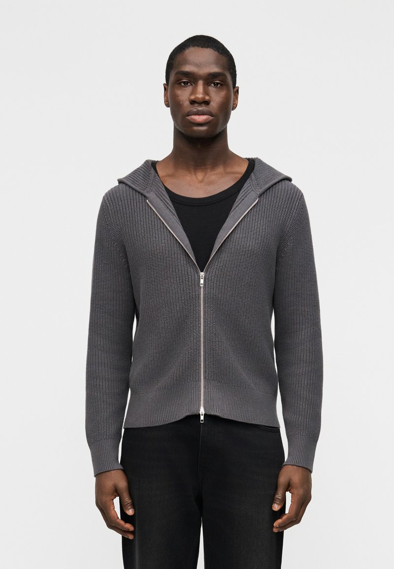 Άνδρας που φοράει γκρι πλεκτό φούτ ερ με φερμουάρ πάνω από μαύρο κοντομάνικο μπλουζάκι και μαύρο παντελόνι, στέκεται μπροστά σε ένα απλό λευκό φόντο.