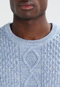 Close-up of a person’s lower face and neck wearing a light blue knit sweater with diamond and cable patterns.
