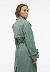 Trench coat verde chiaro con vita speronata, spalline e aletta posteriore. Tessuto morbido con maniche lunghe e dettagli sui polsini.