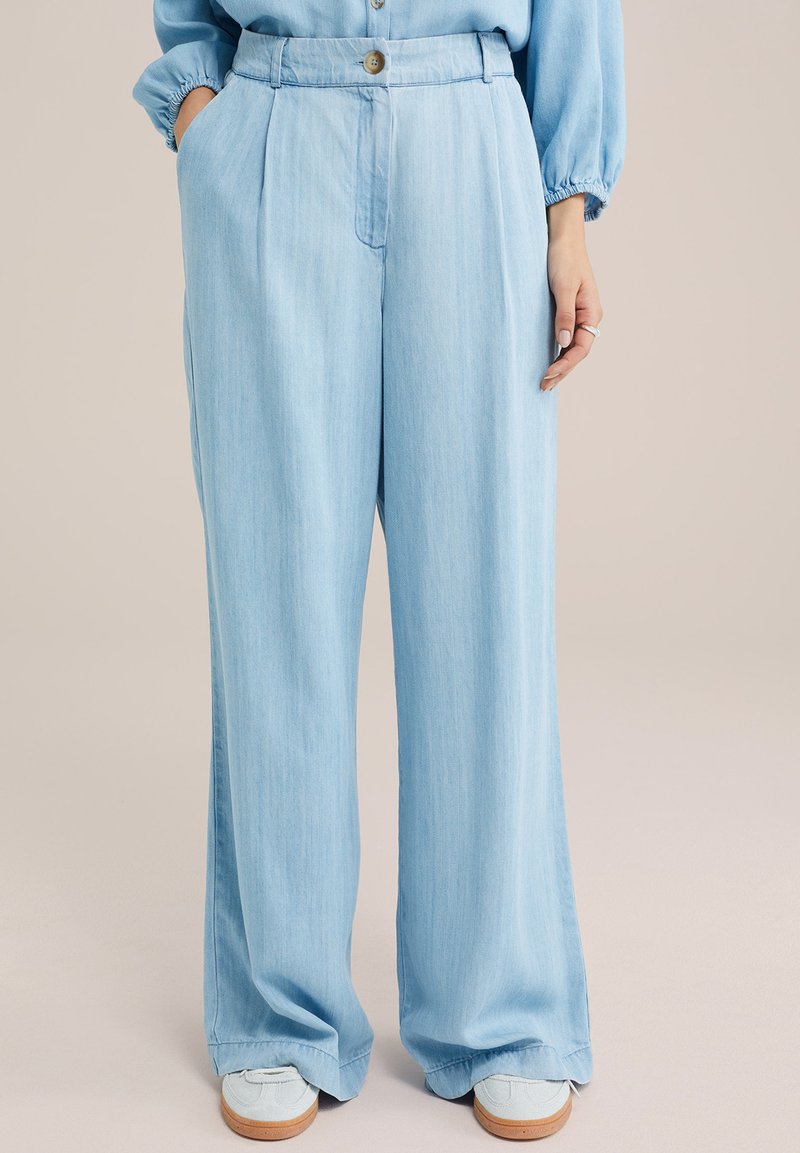 Pantalones de pierna ancha en azul claro, fabricados en un tejido suave, con cinturilla de botón, frente plisado y bolsillos laterales. Combinados con zapatillas de color claro.