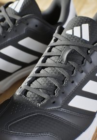 Schwarze Sportschuhe mit grauen Akzenten, drei weißen Streifen, strukturiertem Synthesematerial und flachen Schnürsenkeln. Verfügt über Belüftungsöffnungen für Atmungsaktivität.