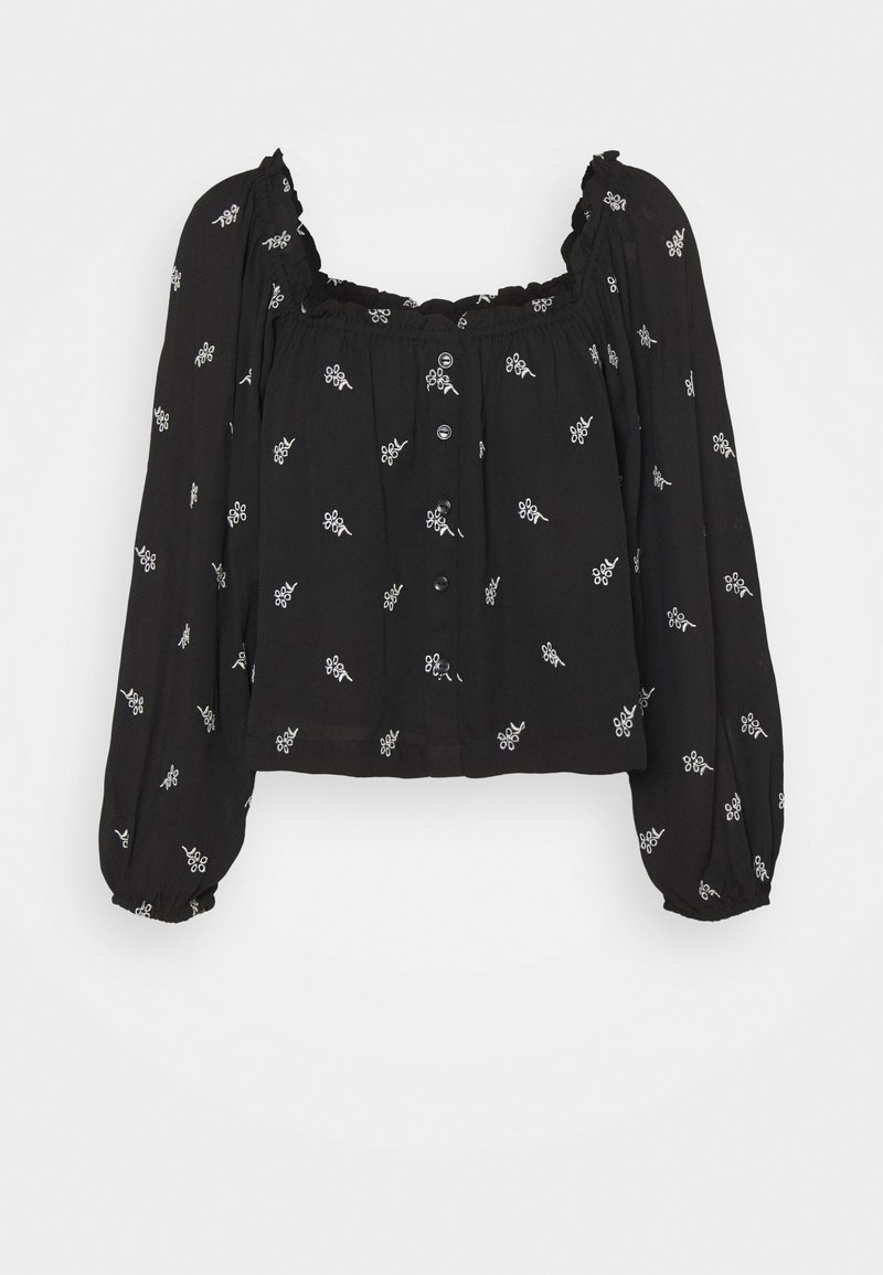 Blouse noire à manches longues avec motifs floraux blancs, boutonnage sur le devant, encolure et poignets élastiqués, et coupe légèrement courte.