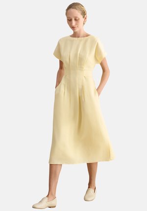 Mujer con un vestido midi amarillo claro con mangas cortas y bolsillos laterales, combinado con zapatos beige tejidos sin cordones, de pie sobre un fondo blanco.