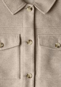 Béžová košile-jacket z vlněné směsi s límcem, čtyřmi knoflíky a dvěma náprsními kapsami. Měkká textura s jemným leskem a kontrastními knoflíky.
