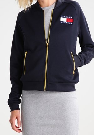 Granatowa kurtka na zamek z złotymi suwakami, dwoma przednimi kieszeniami i naszywką z logo Tommy Jeans na górnej części klatki piersiowej, noszona na szarej sukience.
