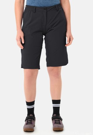 Personne portant un short noir mi-cuisse, des chaussettes noires à rayures blanches, des chaussures de randonnée violettes, et une chemise bleue rentrée dans le short.