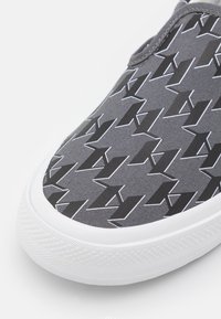 Chaussure grise à enfiler avec semelle blanche avec un motif géométrique de flèches noires et blanches répété sur le tissu supérieur.
