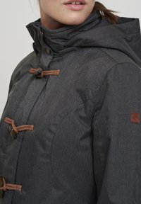 Närbild av en grå huva med toggle-knappar och vävda snoddar som bärs av en person med delvis synlig ansikte.