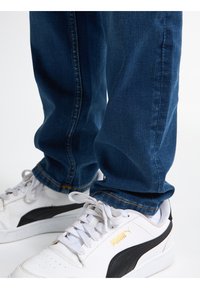 Džínové kalhoty ve tmavě modré barvě se rovný střih, doplněné bílými a černými teniskami s logem, šněrováním a perforovanými detaily.