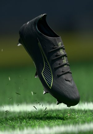 Black soccer cleat with a textured surface, green accents, and visible laces; suspended above green grass, with small debris around.