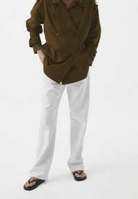 Brązowa podwójnie zapinana kurtka o luźnym kroju z przeszytymi rękawami, w zestawie z białymi spodniami o szerokich nogawkach i czarnymi klapkami.