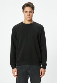 Sudadera negra con cuello redondo, mangas largas y puños acanalados. Confeccionada con un tejido de textura suave, presenta un diseño de corte holgado.