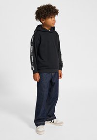 Schwarzer Kapuzenpullover mit Logo-Akzenten an den Ärmeln, kombiniert mit blauen Jeans und beigen Sneakers mit schwarzen Streifen.
