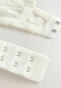 Artimiausias balto audinio liemenėlės sagties užsegimas su metalinėmis kabliukais ir nėriniais šviesiame fone.
