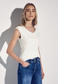 Top bianco senza maniche con scollatura a V abbinato a jeans in denim blu, che mostra una silhouette aderente e una texture morbida.