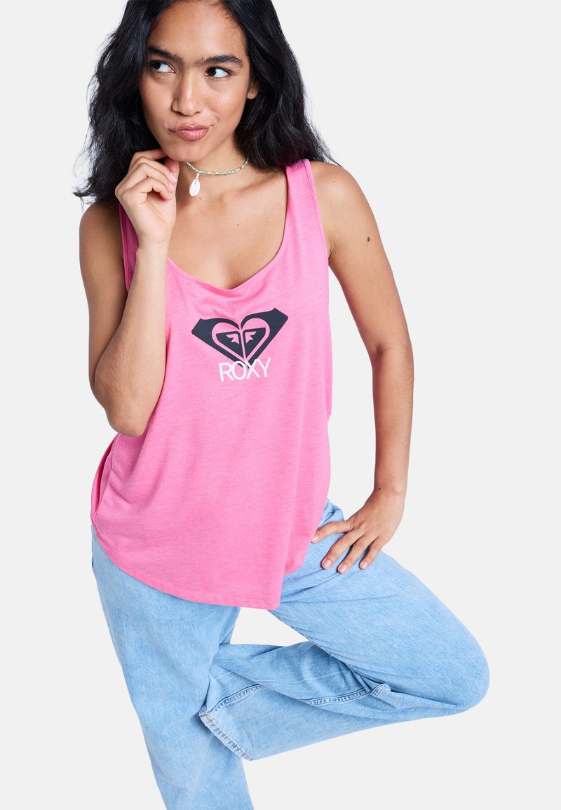 Joven con una camiseta sin mangas rosa de Roxy y jeans azul claro, posando con una rodilla levantada, mano en la cadera y expresión pensativa.