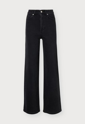 Femme blonde portant un jean noir à jambes larges, un sweat-shirt court gris foncé avec un logo, et des chaussures noires pointues, debout devant un fond uni.