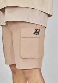 Pantaloni cargo beige in un tessuto resistente, con una tasca laterale e un logo nero. Cuciture piatte e design a taglio dritto.