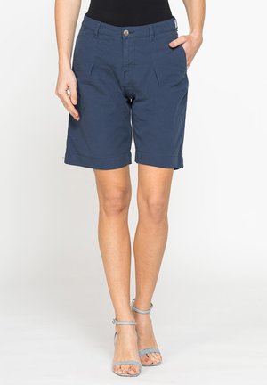 Donna che indossa pantaloncini blu navy fino al ginocchio con pieghe frontali e tasche, abbinati a sandali azzurri con tacco alto e top nero.