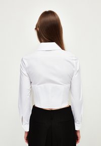 Camicetta bianca corta con maniche lunghe e colletto, con un corpetto aderente e spacchi laterali, abbinata a pantaloni neri a vita alta.