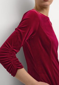 Långärmad, bordeauxklänning i mjukt sammetsmaterial. Har rynkdetaljer på ärmarna och en smal, figurnära silhuett.