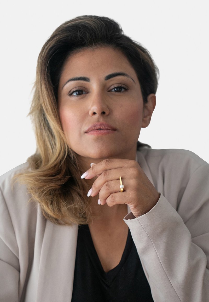 Blazer beige a maniche corte sopra una maglietta nera, con una mano che indossa un anello d'oro con una perla, su uno sfondo bianco semplice.