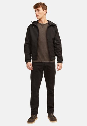 Jonge man met een zwart hoodjack, een bruin overhemd, zwarte broek en bruine schoenen, staand en naar links kijkend tegen een effen achtergrond.