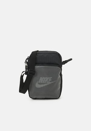 Sac bandoulière compact noir et gris avec une sangle réglable et un logo Nike pixélisé blanc sur la poche avant, sur fond uni.