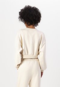 Lys beige kort sweatshirt med puffy ermer, ribbestrikkede mansjetter og bredt liv. Prægede logo detaljer på bagsiden. Glat tekstur.