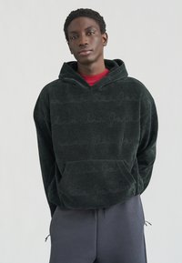 Σκούρο πράσινο φούτερ με κουκούλα από απαλό υλικό με υφή, με μοτίβο λογότυπου στον ίδιο τόνο και μπροστινή τσέπη· φορεμένο πάνω από κόκκινο πουκάμισο.