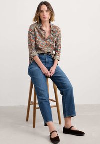 Blus med blommönster i brunt, med livfulla färger; högmidjade blå jeans; bruna platta skor; sitter på en träpall.