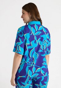 Blusa a maniche corte in blu e viola, con un motivo floreale e accenti verde chiaro. Tessuto morbido, vestibilità comoda e colletto.