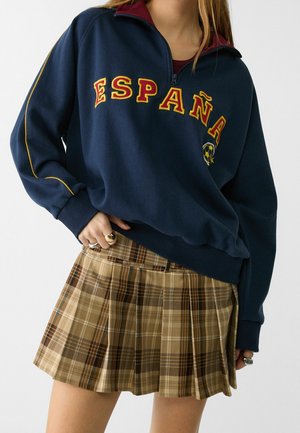 Personne portant un sweat-shirt bleu marine avec le texte "España" et un petit ballon de football, associé à une jupe plissée marron à carreaux.