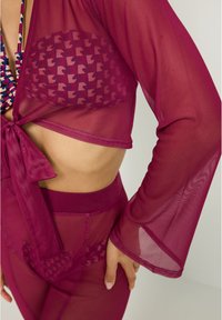 Cubre que es transparente en magenta con mangas largas y amplias, atado en la parte delantera, revelando un sujetador de estampado y unas braguitas de tiro alto a juego por debajo.